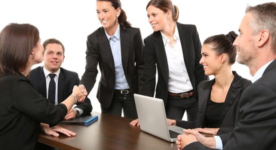 Réunion professionnelle avec six personnes, une femme serrant la main d'une autre, et des participants souriants autour d'une table avec des ordinateurs, symbolisant la collaboration et le soutien professionnel.